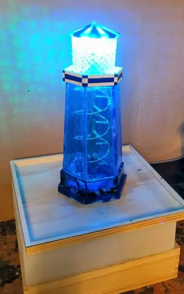 a resin lighthouse with a blue green LED light inside the lens room, a black and white painted widow's walk surrounds the bright light, and a spiral staircase ascends to the light through the middle of the blue resin walls in the shape of a hexagon and which sit on a blue resin base containing rocks at the ocean level